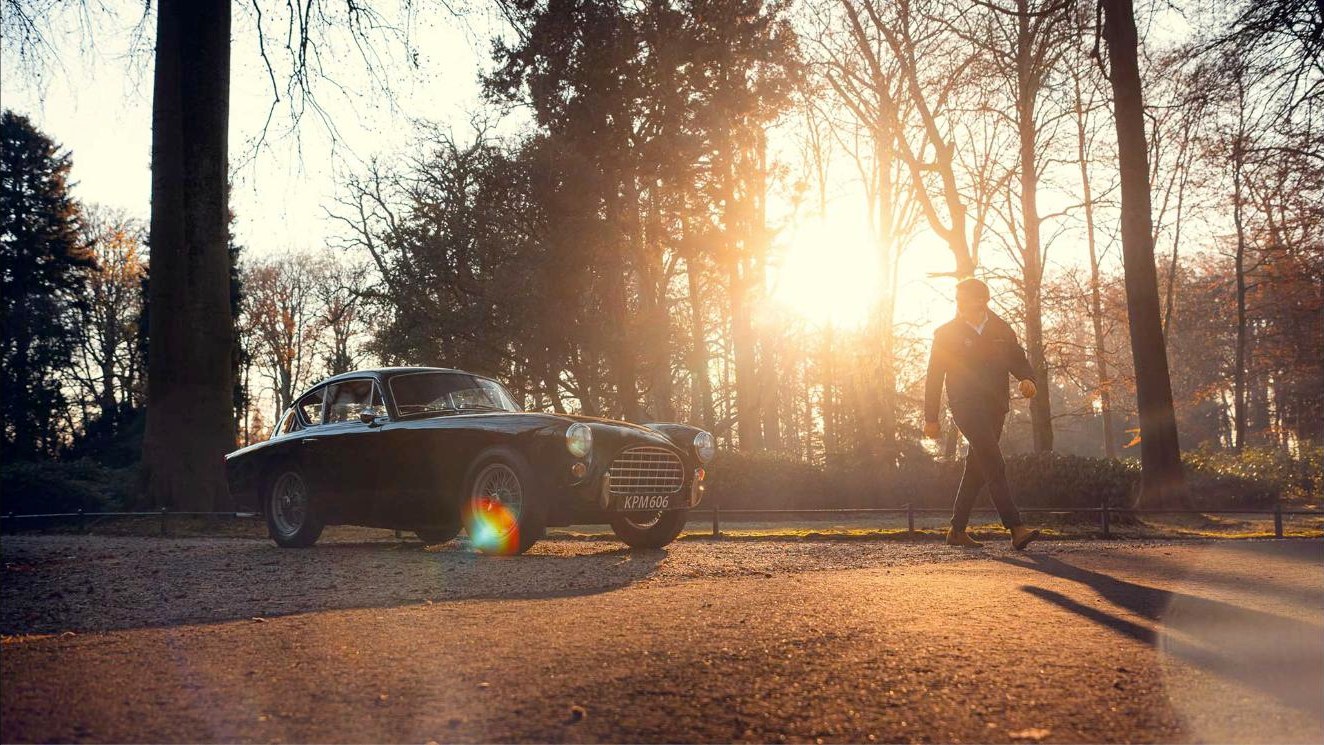 Man loopt in herfstlicht naar een klassieke sportwagen op een bosweg